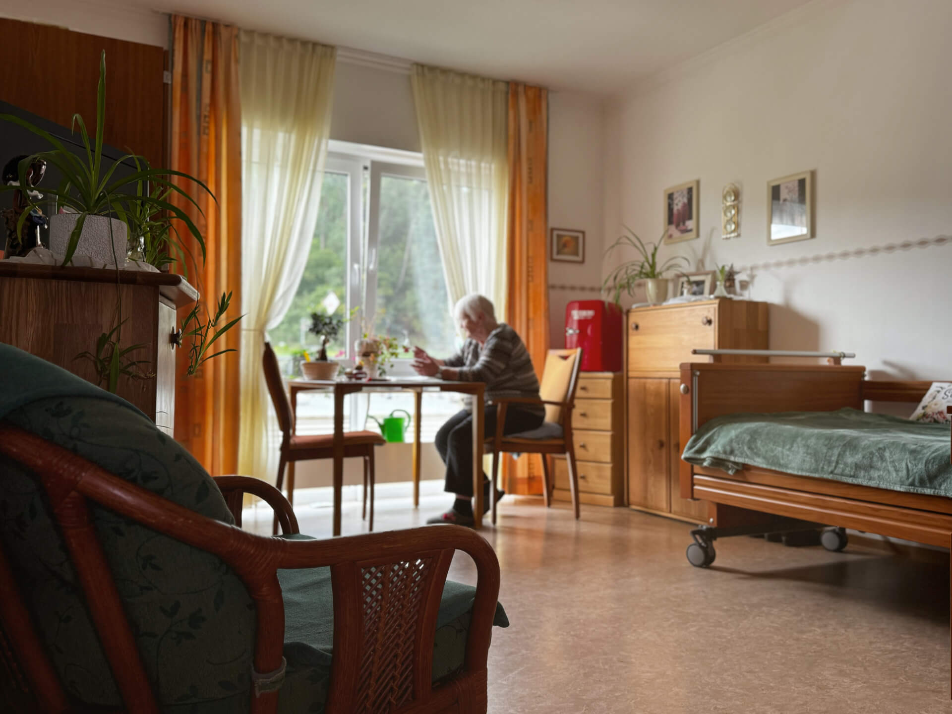 Bewohnerin an einem Tisch sitzend. Bett, Kommode und frische Blumen.